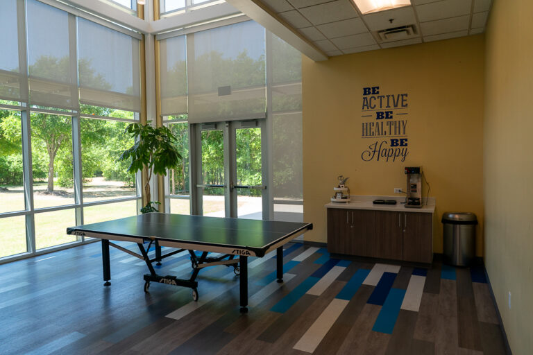A ping pong table is set up in a bright room with large windows, a plant, a coffee station, and a wall quote that says, "Be active, be healthy, be happy.
