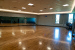 An empty exercise studio with wooden floors, mirrored wall, stacked equipment in the corner, ceiling lights, and large windows letting in natural light.