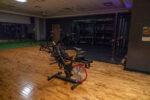 A dimly lit gym room with an exercise bike in the foreground and various strength training equipment in the background on a polished wooden floor.