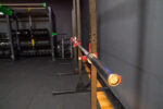 A barbell rests on a rack in an empty gym, with weight plates and other equipment visible in the background.