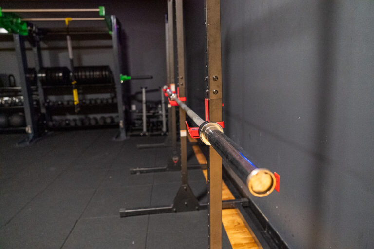 A barbell rests on a rack in an empty gym, with weight plates and other equipment visible in the background.