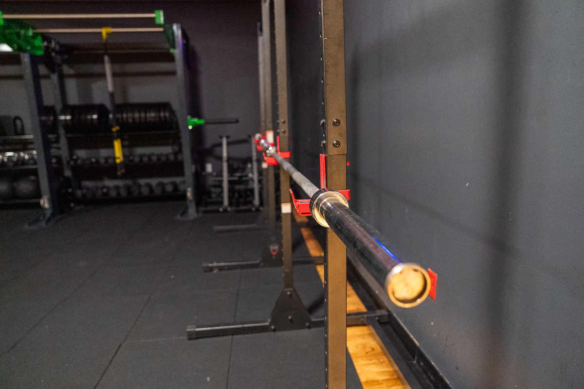A barbell rests on a rack in an empty gym, with weight plates and other equipment visible in the background.