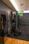 A gym corner with barbells, weight plates, resistance bands, and a squat rack on a rubber floor next to a digital clock.