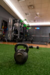 A black kettlebell sits on artificial turf in a gym, with exercise equipment, weights, and racks visible in the background.