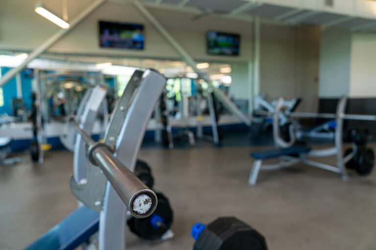Close-up of an empty barbell on a rack in a gym, with various exercise equipment and TV screens visible in the background.