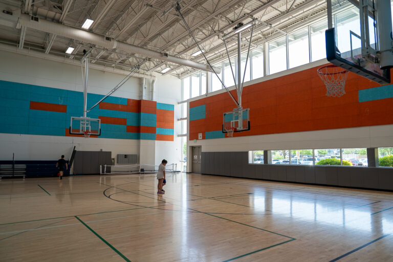 A person stands on a basketball court in a well-lit gymnasium with high ceilings and colorful wall panels.