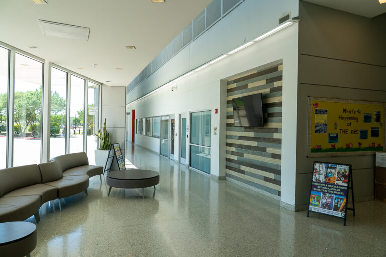 Modern lobby area with curved seating, round tables, large windows, a bulletin board, wall-mounted TV, and polished floors.