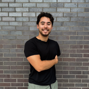 A man in a black t-shirt and green pants stands in front of a gray brick wall, smiling with his right arm folded across his body, representing our team’s friendly spirit.