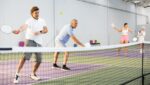 Four adults enjoy indoor pickleball at The Rec at The Lakefront™, with two men in the foreground returning the ball while two women stand ready in the background.