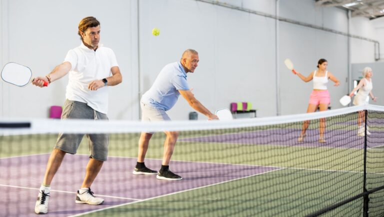 Four adults enjoy indoor pickleball at The Rec at The Lakefront™, with two men in the foreground returning the ball while two women stand ready in the background.