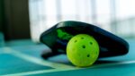 A close-up of a pickleball paddle and a perforated yellow pickleball resting on the green court at The Rec at The Lakefront™, perfect for Indoor Pickleball enthusiasts.