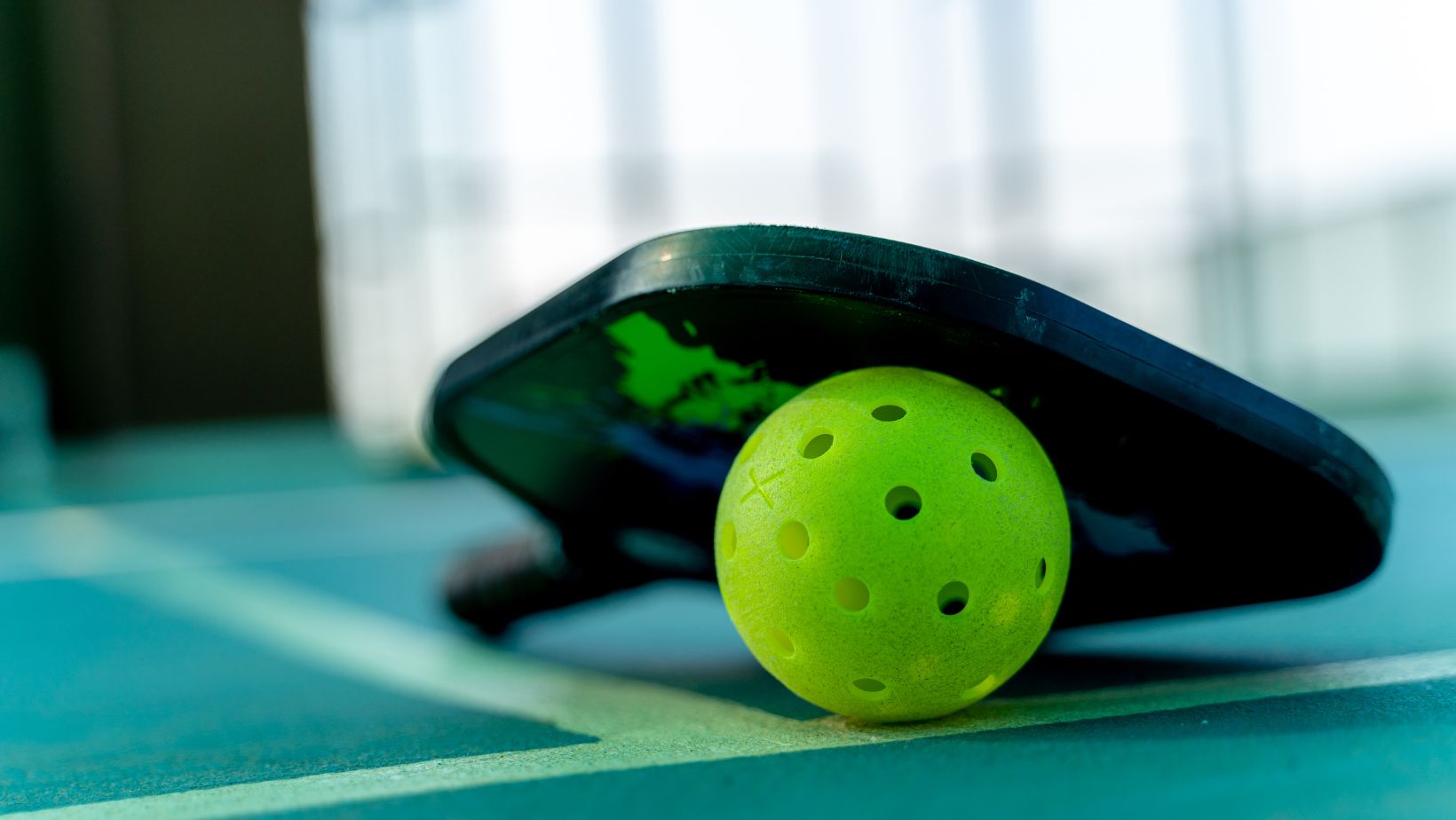 A close-up of a pickleball paddle and a perforated yellow pickleball resting on the green court at The Rec at The Lakefront™, perfect for Indoor Pickleball enthusiasts.