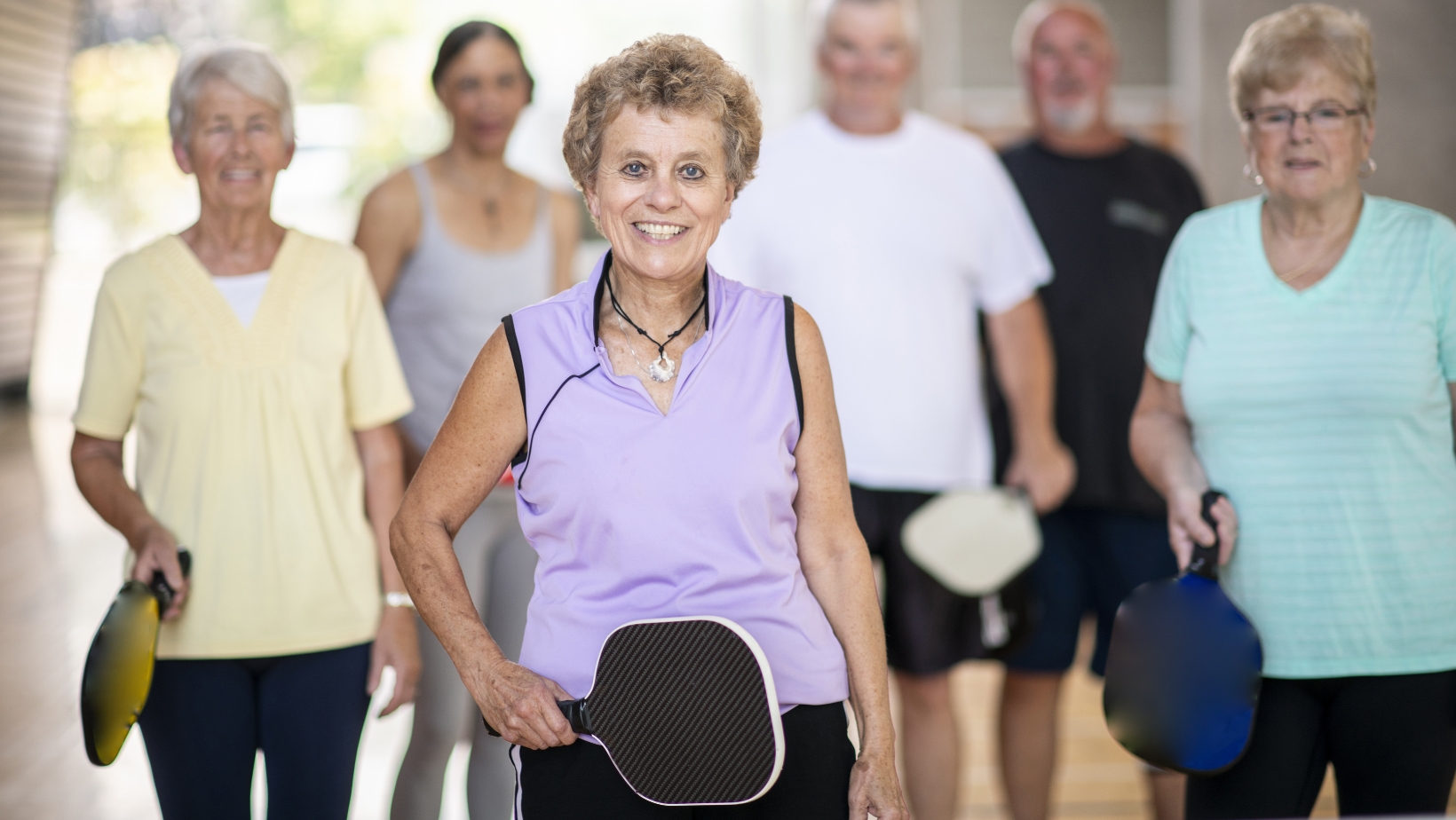 A group of older adults stands indoors, holding pickleball paddles, with one woman in a lavender top smiling at the front—ready to dive into The Rec™ Beginner's Guide and learn about Pickleball Scoring together.