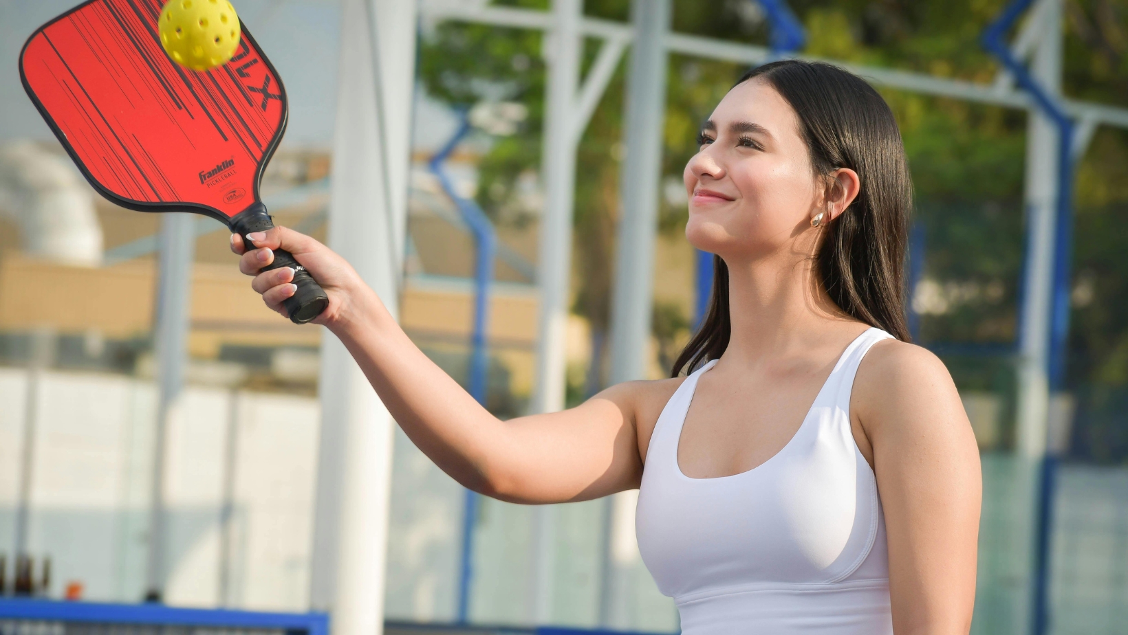 A woman in a white tank top plays pickleball outdoors, holding a red paddle and about to hit a yellow ball, perfectly illustrating tips from The Rec™ Beginner's Guide and the basics of pickleball scoring.