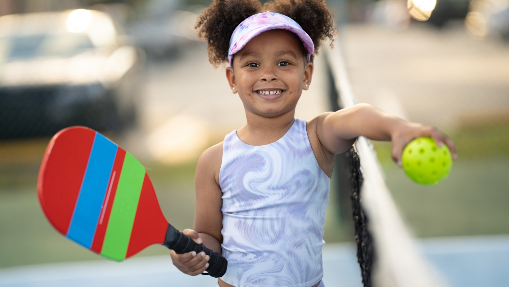 Young girl standing on a tennis court, holding a colorful paddle in one hand and a yellow perforated ball in the other, smiling at the camera—ready to try Pickleball Scoring from The Rec™ Beginner's Guide.
