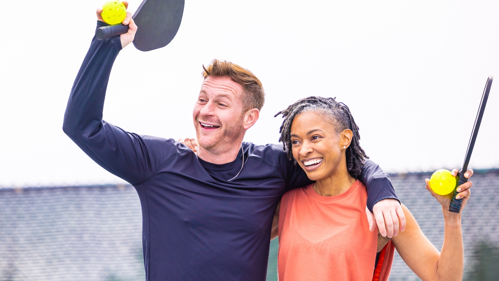 Two people stand side by side outdoors, smiling and holding pickleball paddles with yellow balls, looking happy after a game inspired by The Rec™ Beginner's Guide to Pickleball Scoring.