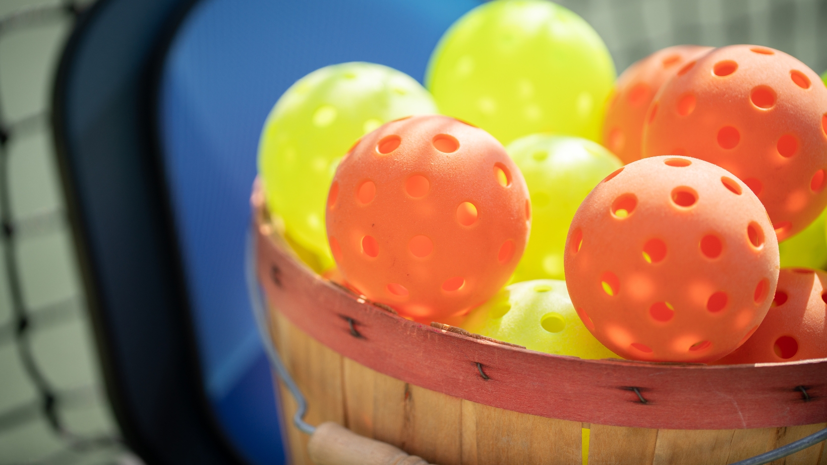 A wooden basket filled with yellow and orange perforated plastic balls, commonly used for playing pickleball, sits near a net—perfect for practicing after reading The Rec™ Beginner's Guide or brushing up on Pickleball Scoring.