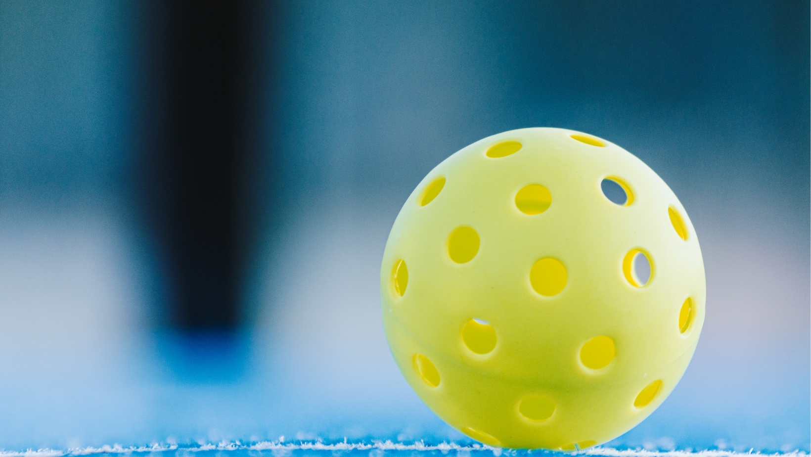 A close-up of a yellow perforated plastic ball resting on a blue surface, with a blurred background—perfect for illustrating Pickleball Scoring from The Rec™ Beginner's Guide.
