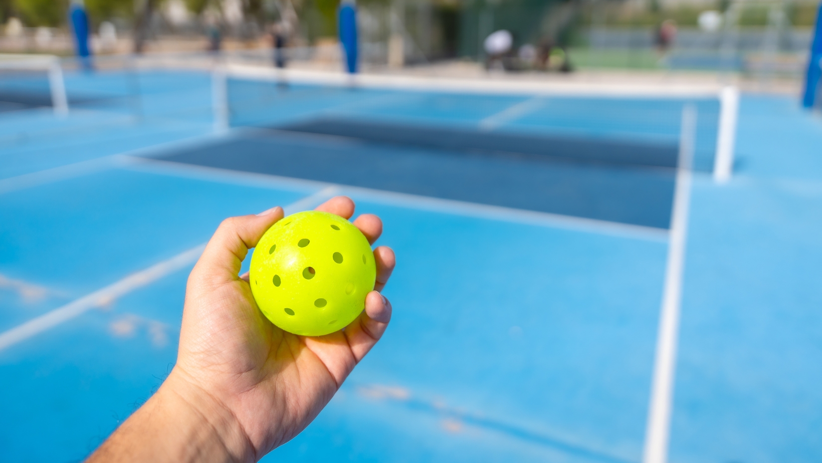 A person holds a yellow perforated pickleball in their hand on an outdoor blue court, ready to practice Pickleball Scoring from The Rec™ Beginner's Guide.
